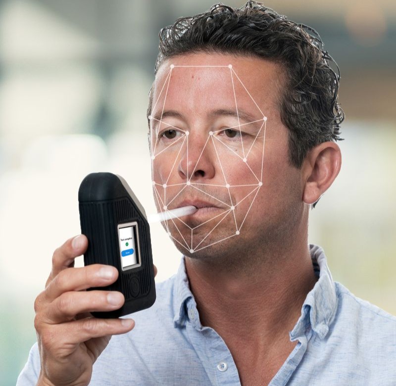 Man using BI SoberTech alcohol monitoring device, blowing into straw with facial biometrics overlay verifying client identity during alcohol test.