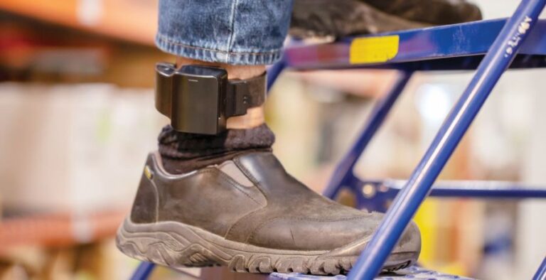 A man uses a step ladder at work. His ankle-worn electronic monitoring device is shown.